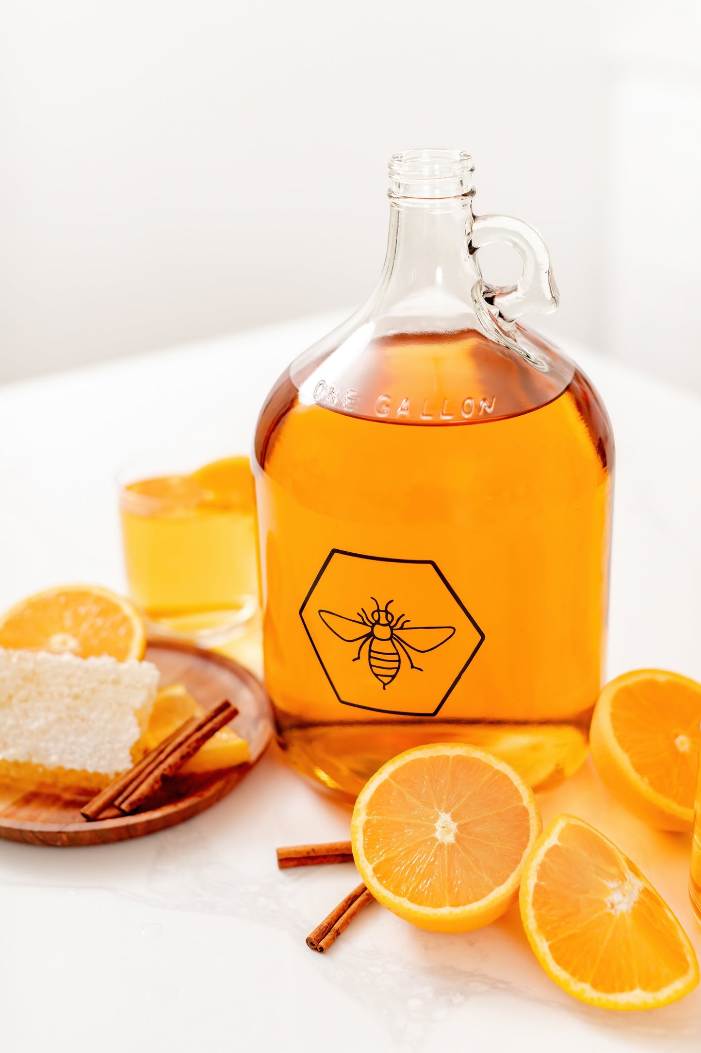 Glass bottle of orange juice with a honeycomb and bee logo, surrounded by oranges and cinnamon sticks on a white surface.
