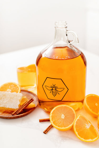 Glass bottle of orange juice with a honeycomb and bee logo, surrounded by oranges and cinnamon sticks on a white surface.